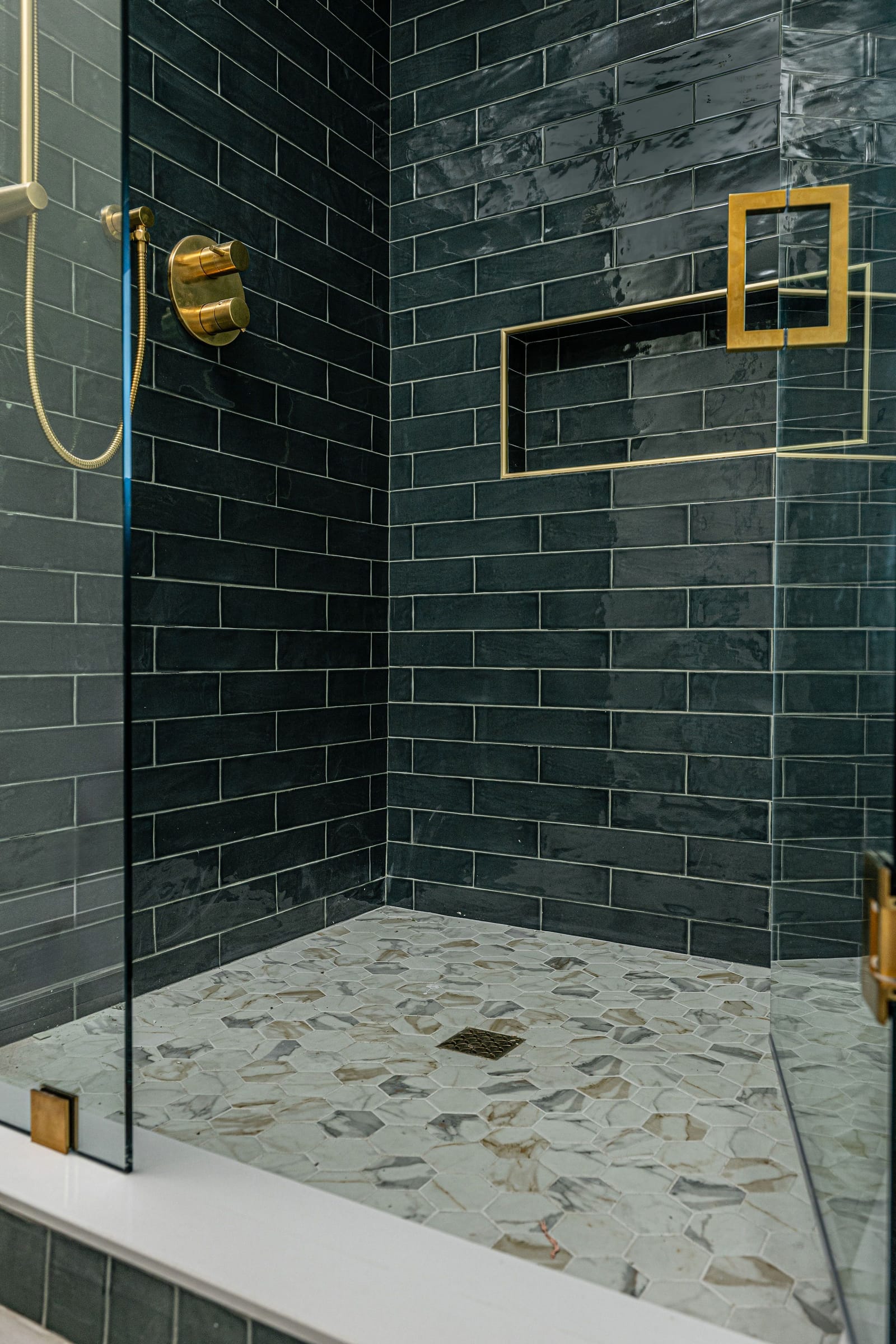 A shower tiled in glossy emerald subway tile, with a brass-trimmed niche, brass fixtures and a marble hexagon mosaic floor