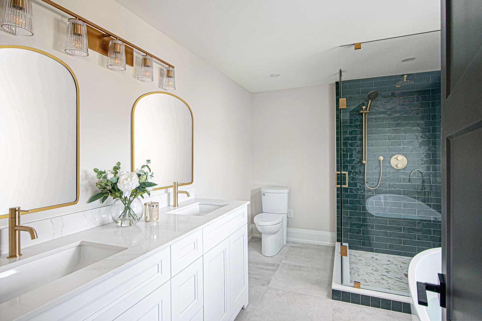 A primary ensuite with a white double vanity, two arched brass mirrors, a ribbed-glass bar light and a glass shower in emerald tile