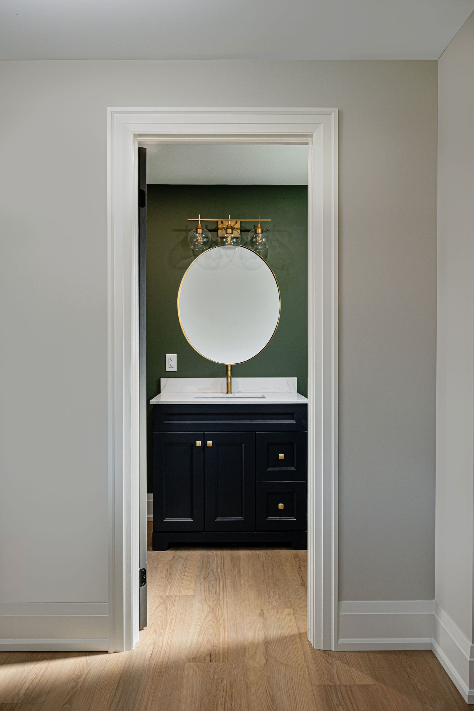 Through a doorway, a bathroom with dark green walls, a navy vanity with brass knobs and an oval brass mirror