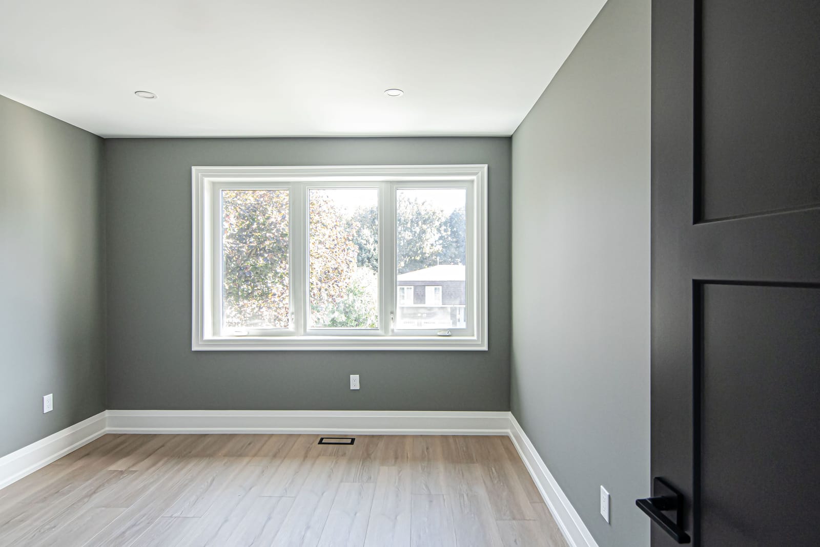 An empty bedroom painted sage grey-green with a wide three-panel window and a dark grey door