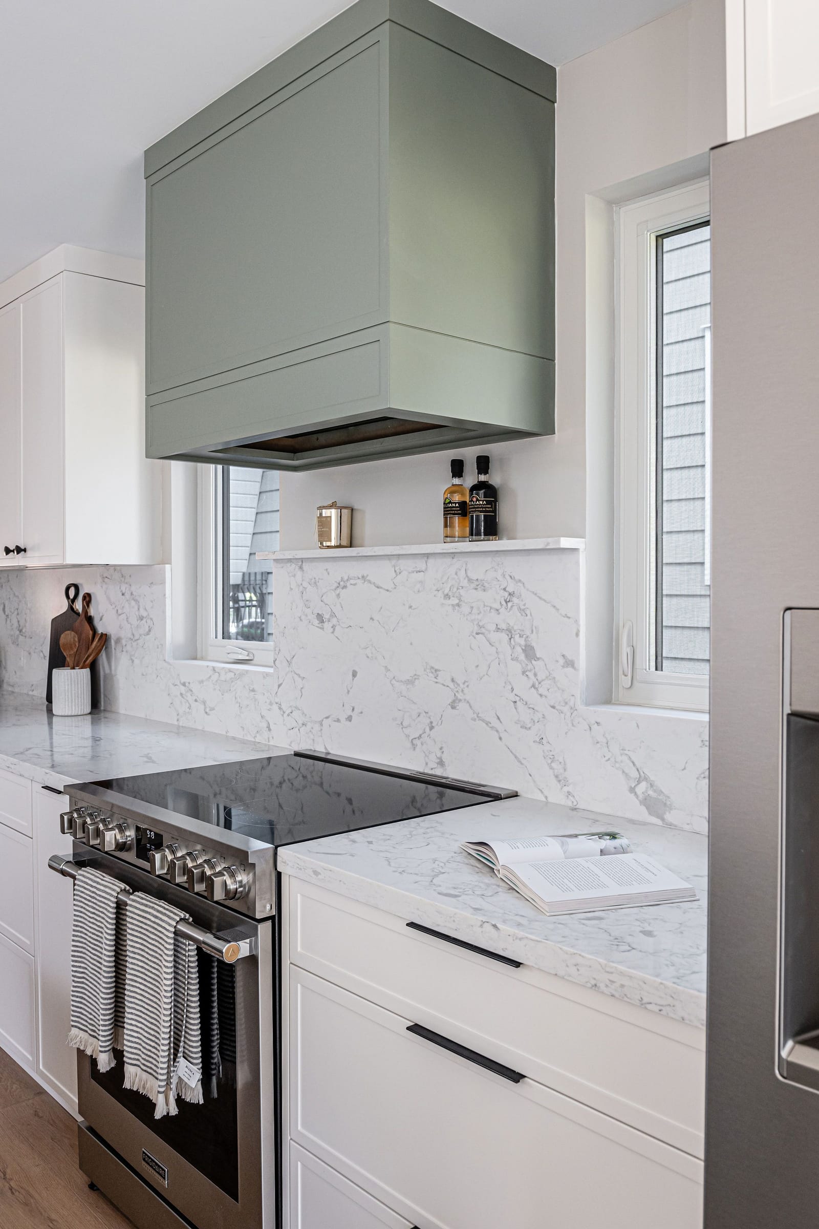 A green box range hood over a stainless range, with a white quartz slab backsplash and a ledge, next to a window
