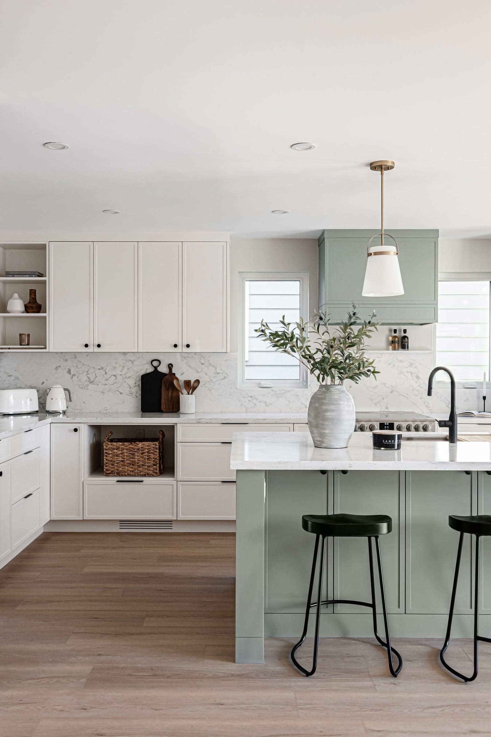 A sage-green island with a white quartz top and black faucet, in front of white cabinets, a marble-look backsplash and a green hood