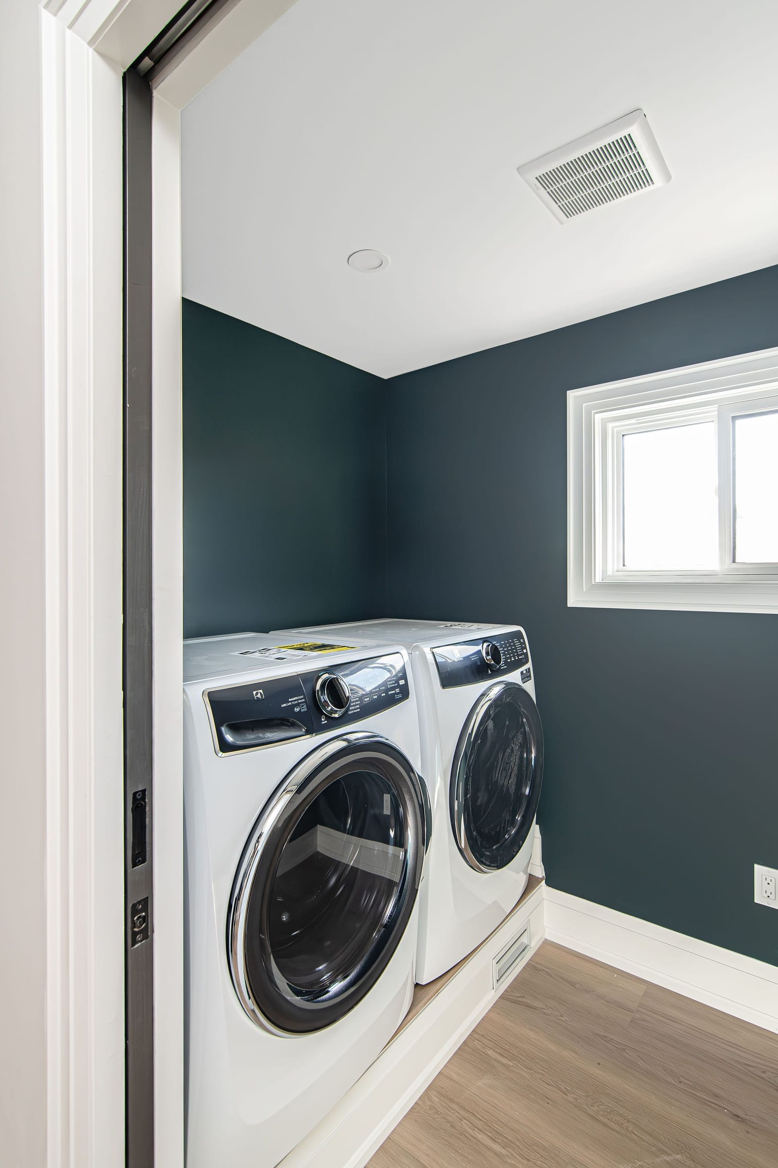 A laundry room with dark teal walls, a white front-load washer and dryer on a raised platform and a small window