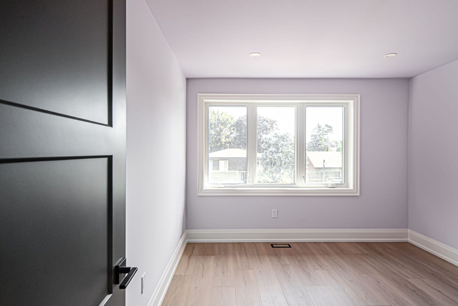 An empty bedroom painted lilac with a wide three-panel window and an oak-look floor
