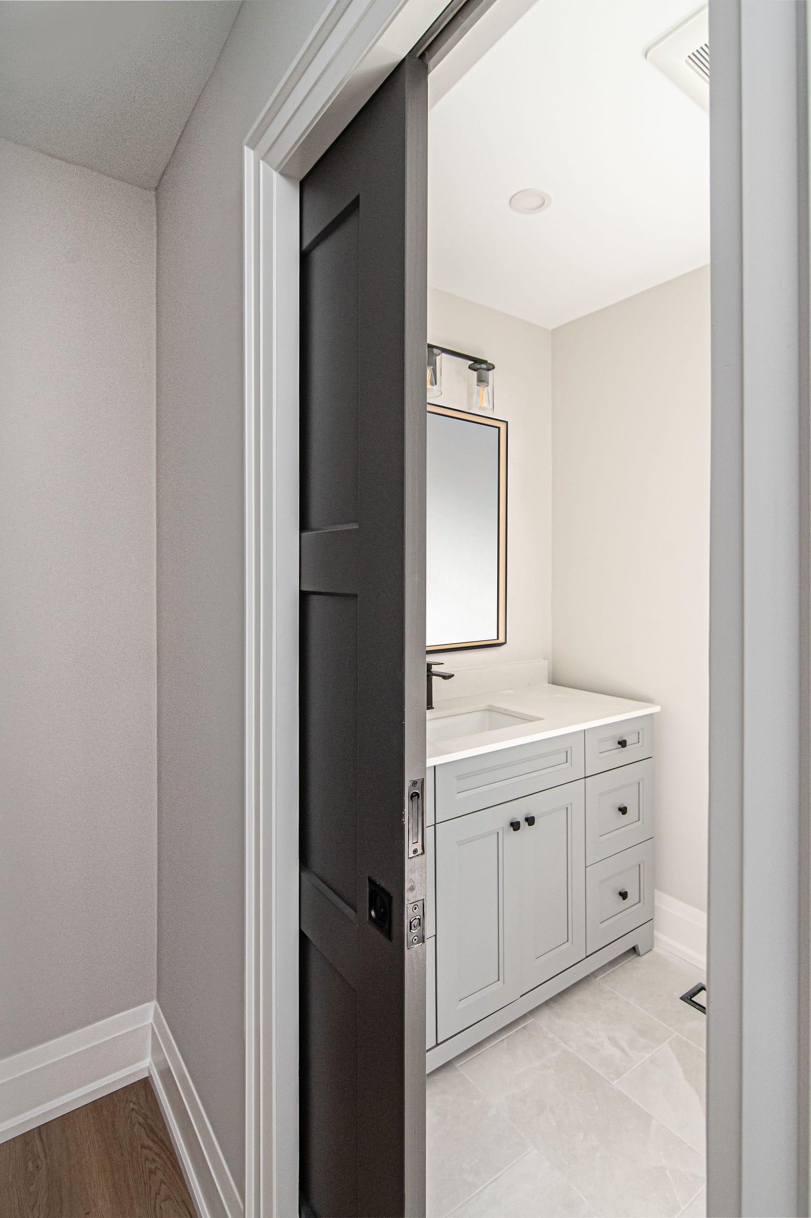 A dark grey pocket door slid open into a small bathroom with a white vanity and a brass-framed mirror