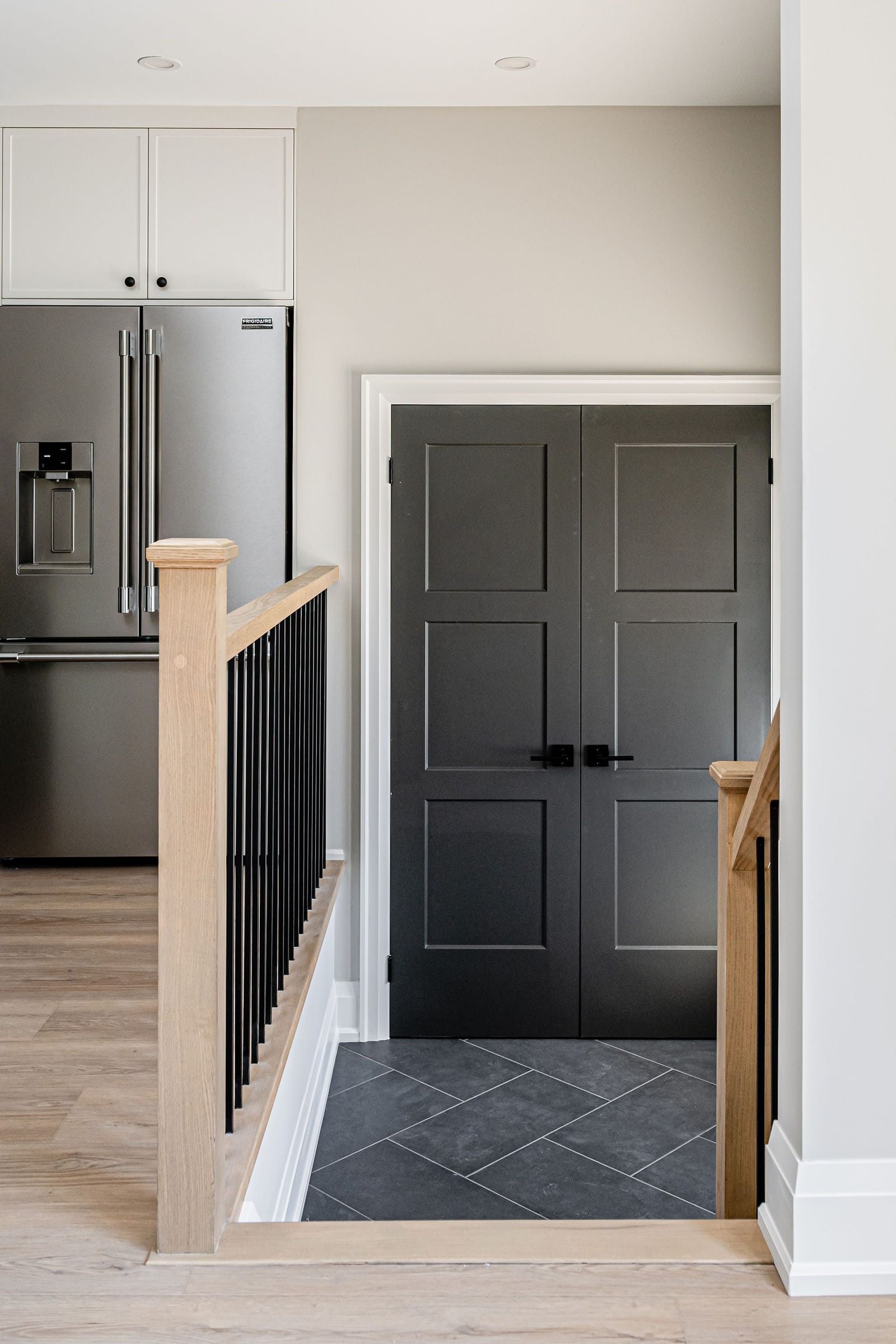 An oak stair rail with black spindles beside dark grey double doors over a slate-look tile landing