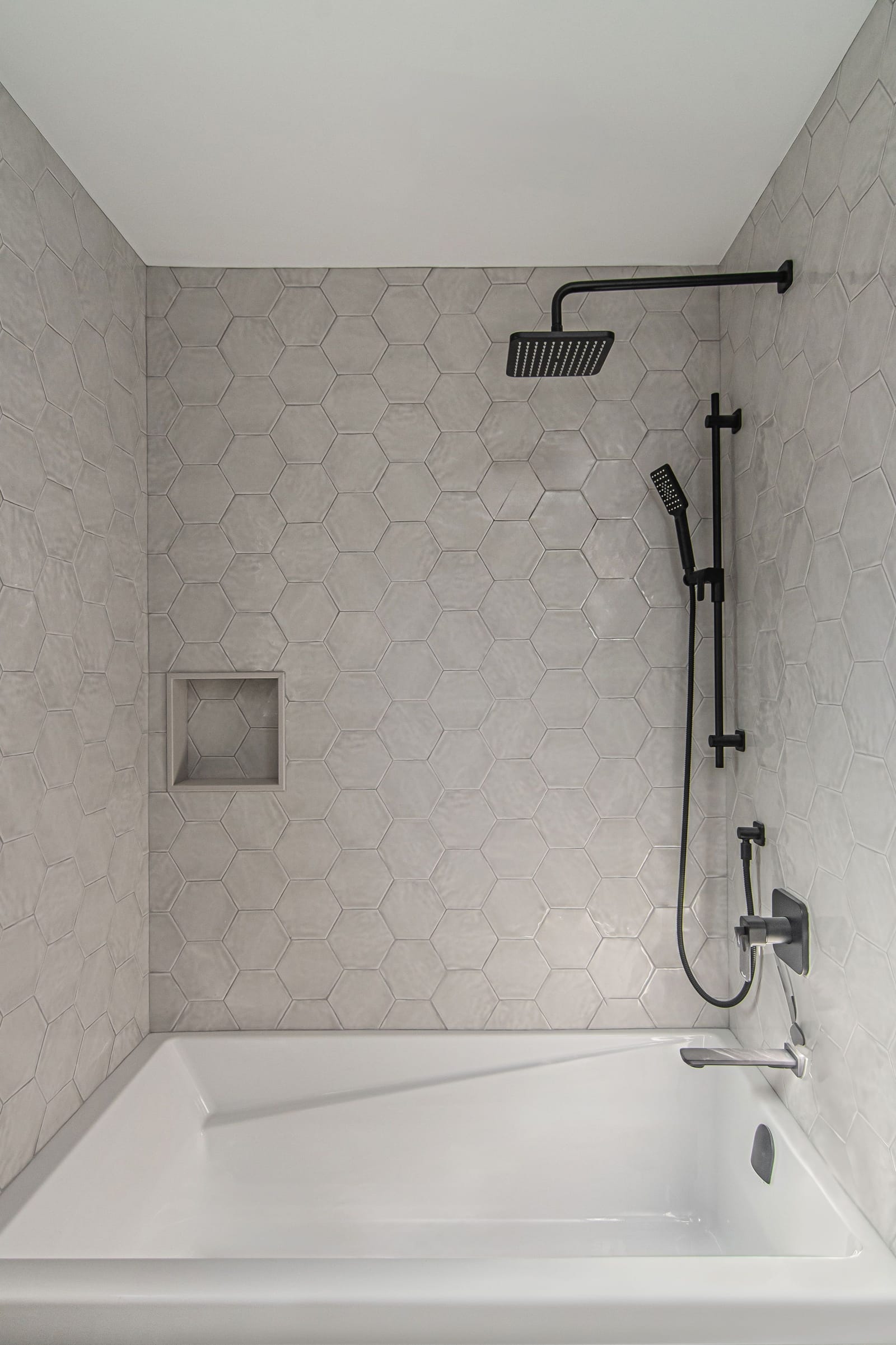 A tub surround in pale grey hexagon wall tile with a square niche and a black rain shower and hand shower
