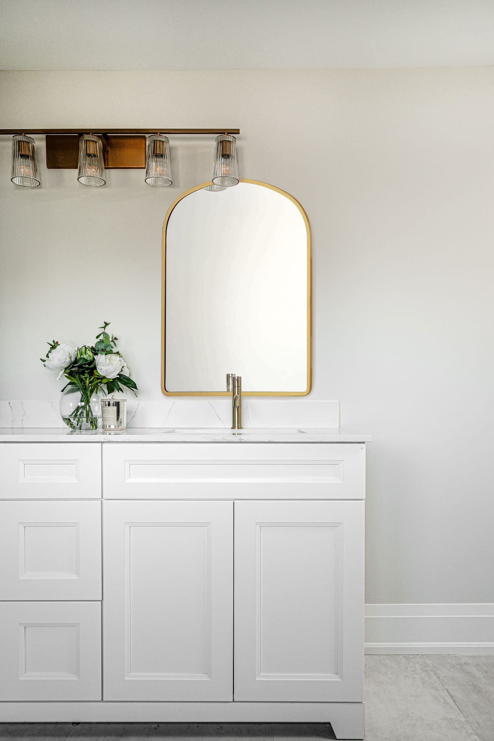 A white shaker vanity with a brass faucet under an arched brass mirror and a four-light bar fixture