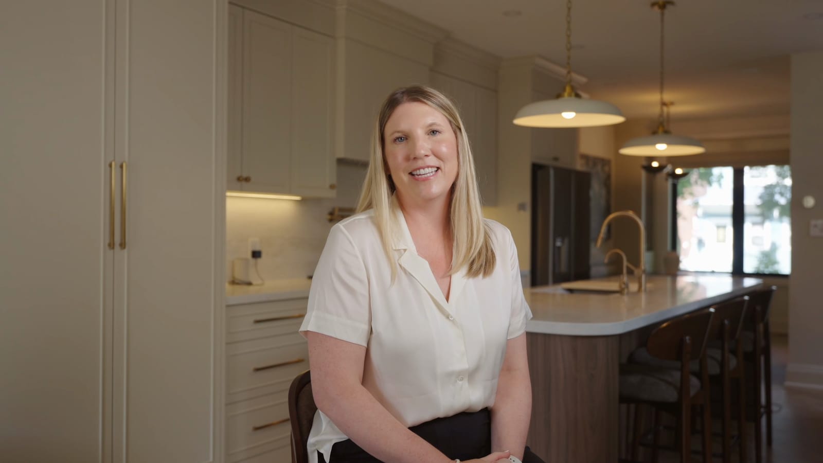 A Greenwood homeowner seated in her finished kitchen, smiling, with cream cabinets, a walnut-sided island and brass pendant lights behind her