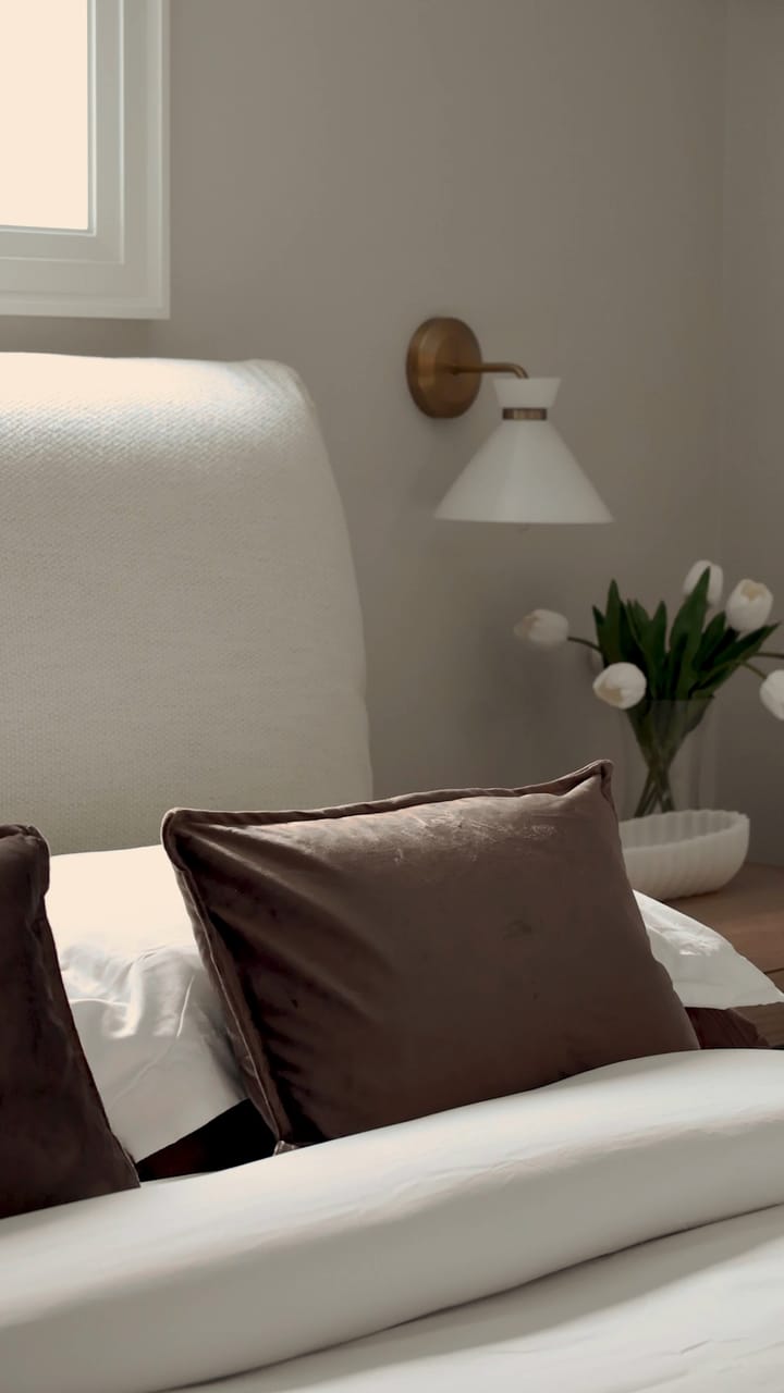 A basement bedroom with a white upholstered bed, brown velvet pillows, a brass and white wall sconce and white tulips on the nightstand